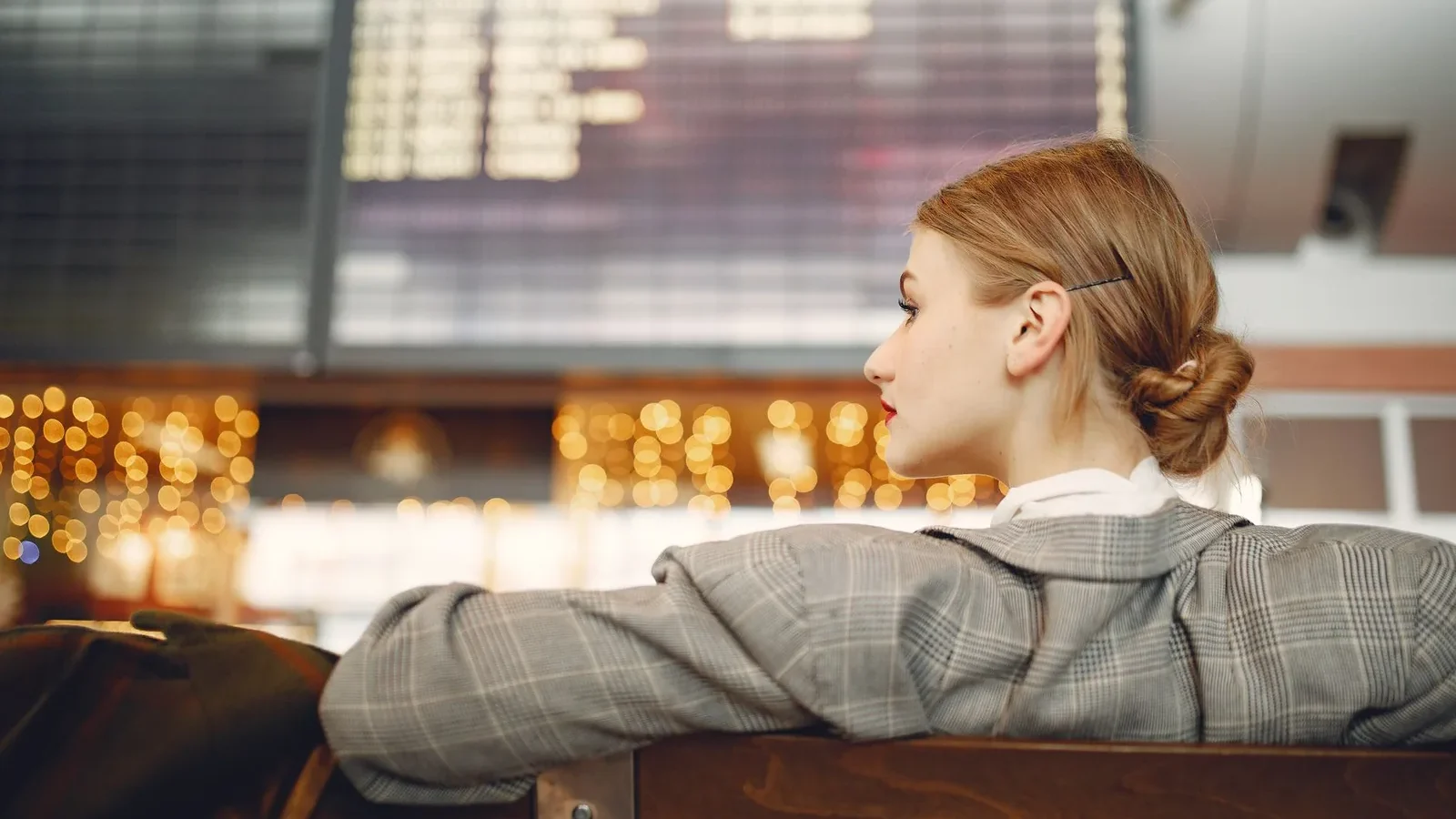 Pensive female designer in checkered jacket waiting for flight near departure board looking away in modern airport in evening.