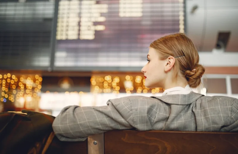 Pensive female designer in checkered jacket waiting for flight near departure board looking away in modern airport in evening.