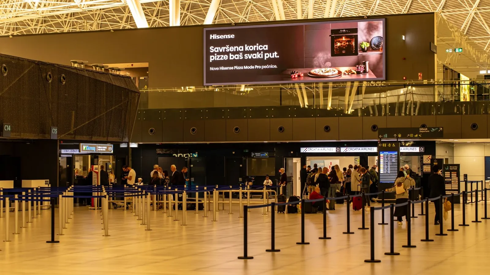 Travelers check in at Zagreb Airport with large advertising screen above.