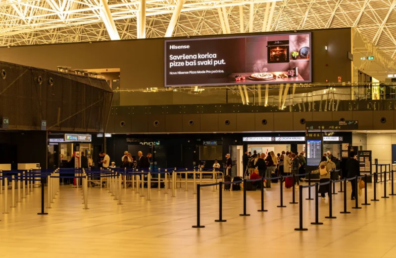 Travelers check in at Zagreb Airport with large advertising screen above.