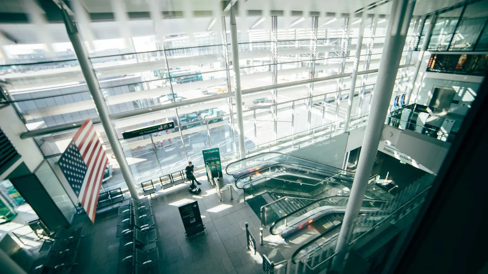 A bright, spacious airport terminal featuring expansive glass walls, escalators, and American flag.