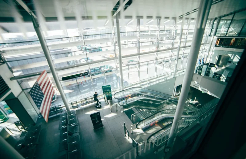 A bright, spacious airport terminal featuring expansive glass walls, escalators, and American flag.