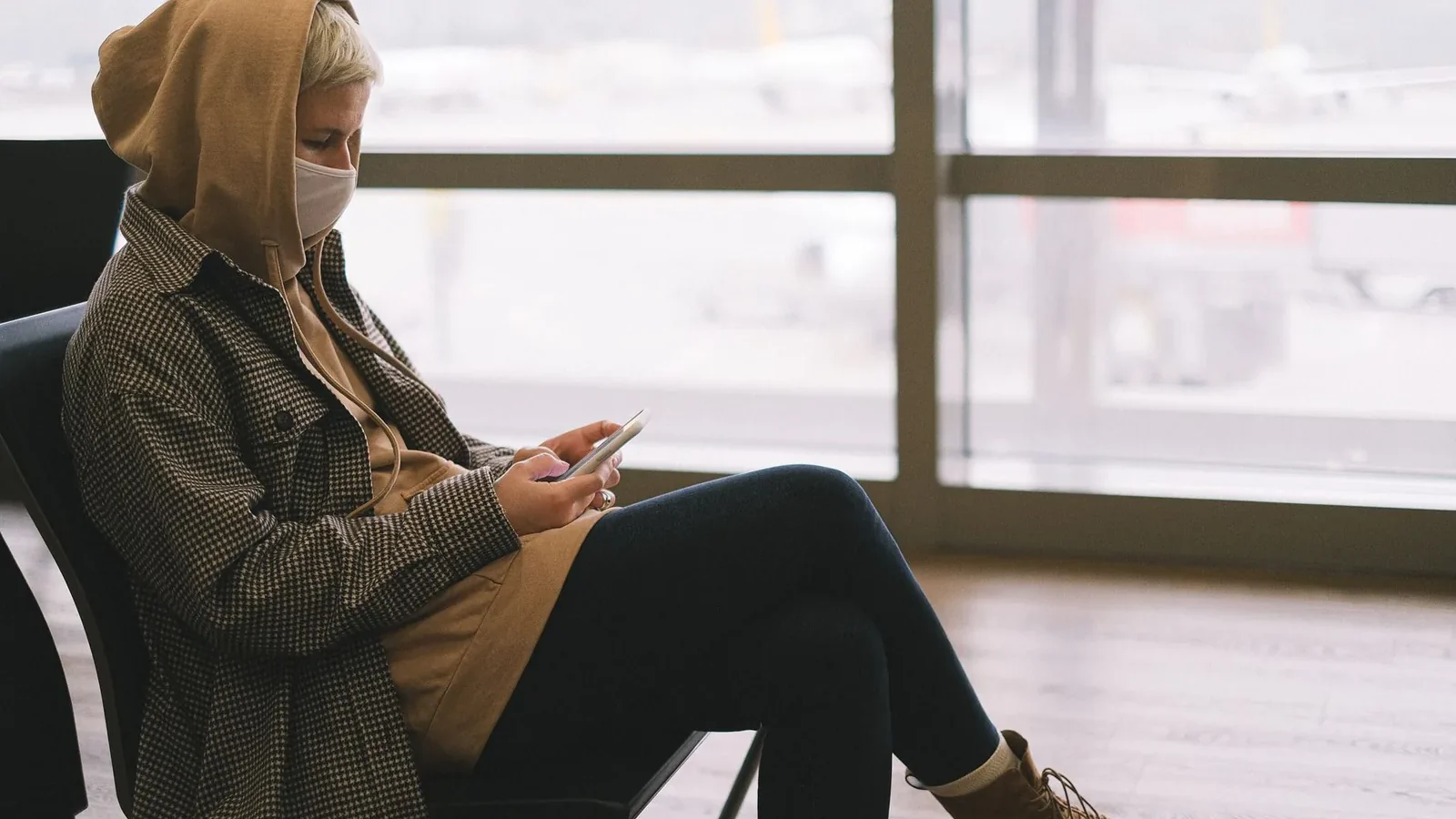 A woman in a hoodie and face mask uses a smartphone while waiting at an airport terminal.