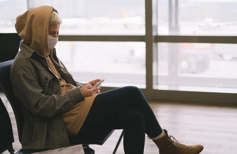 A woman in a hoodie and face mask uses a smartphone while waiting at an airport terminal.