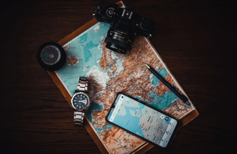 Top view of travel essentials including a map, camera, phone, watch, and lens on a wooden surface.