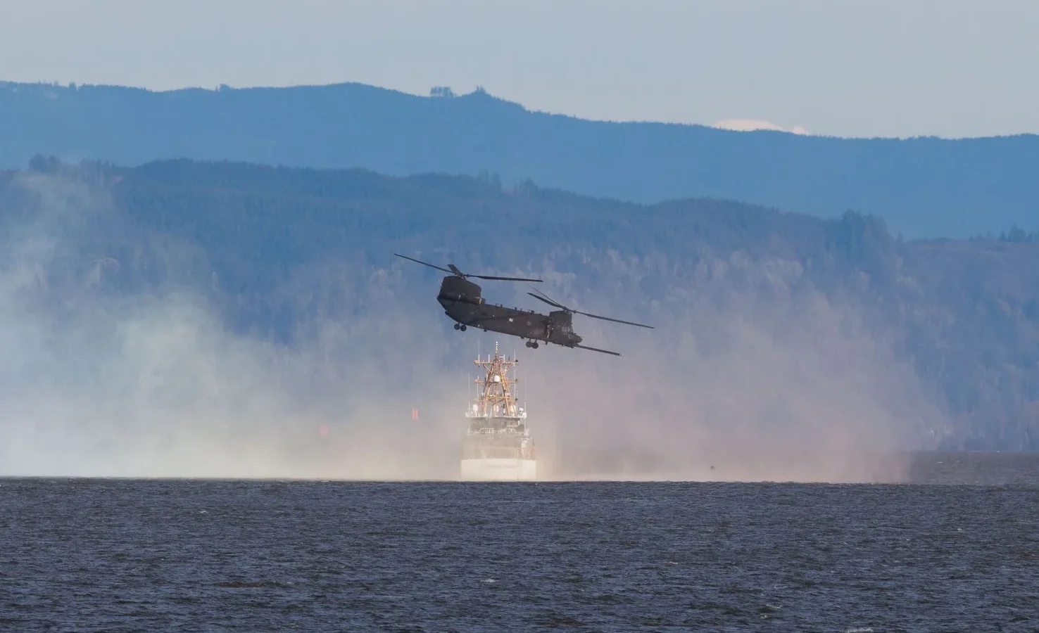 A helicopter flies over a body of water with mountains in the background, creating a dramatic scene.