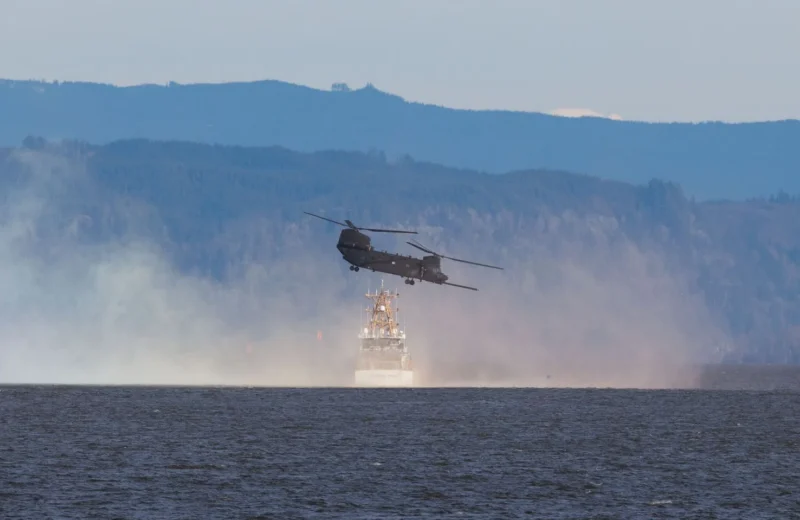 A helicopter flies over a body of water with mountains in the background, creating a dramatic scene.