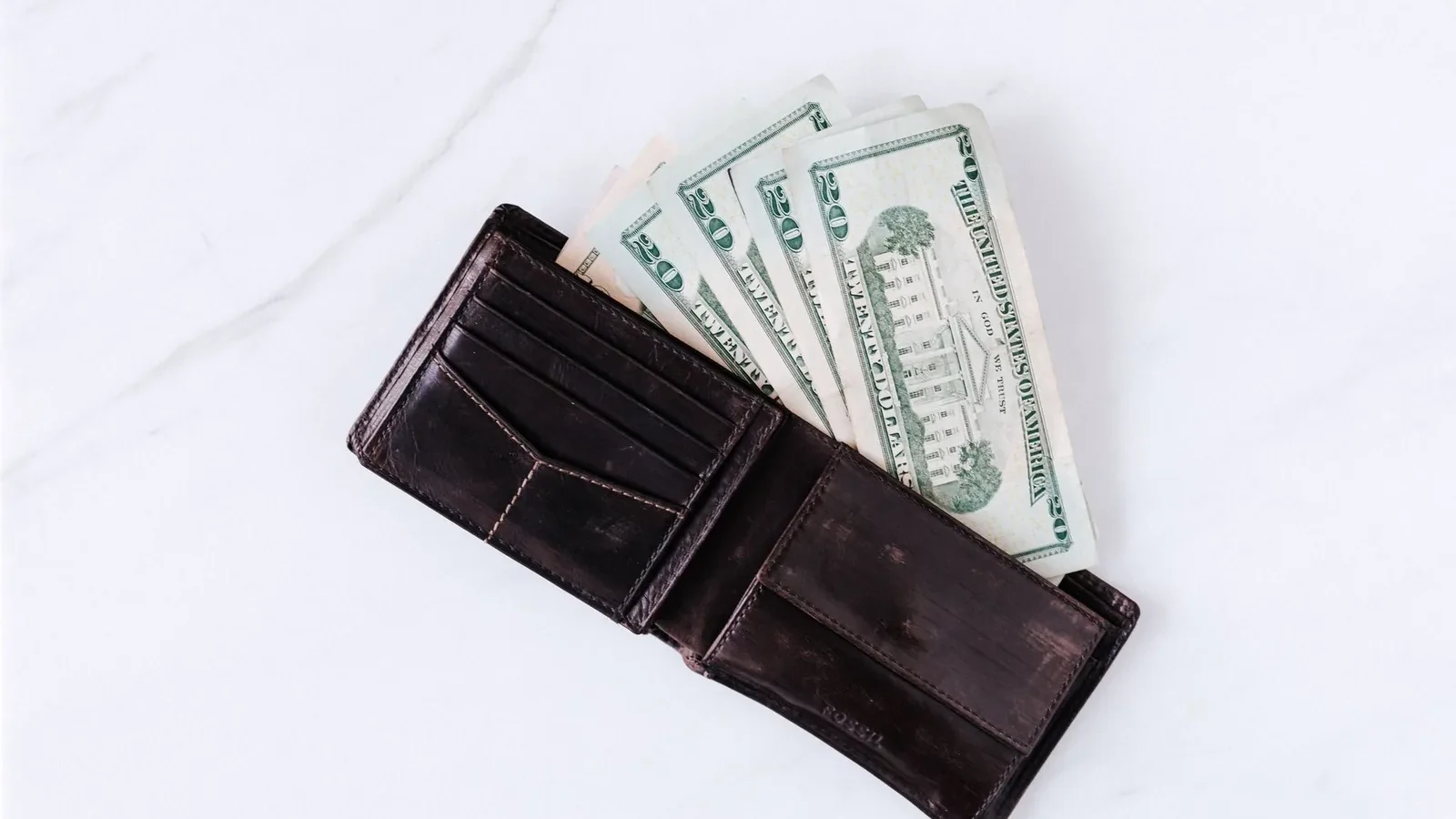 From above of black leather wallet with pockets filled with heap of dollar banknotes on white table.