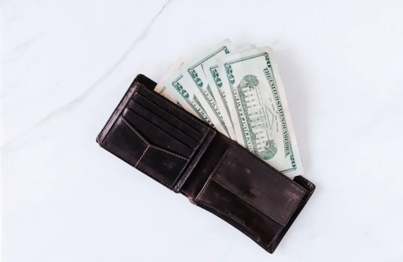 From above of black leather wallet with pockets filled with heap of dollar banknotes on white table.