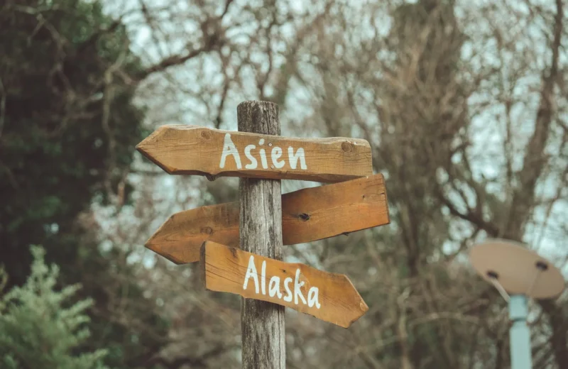 Rustic wooden directional signposts pointing towards Asien and Alaska in a natural outdoor setting.