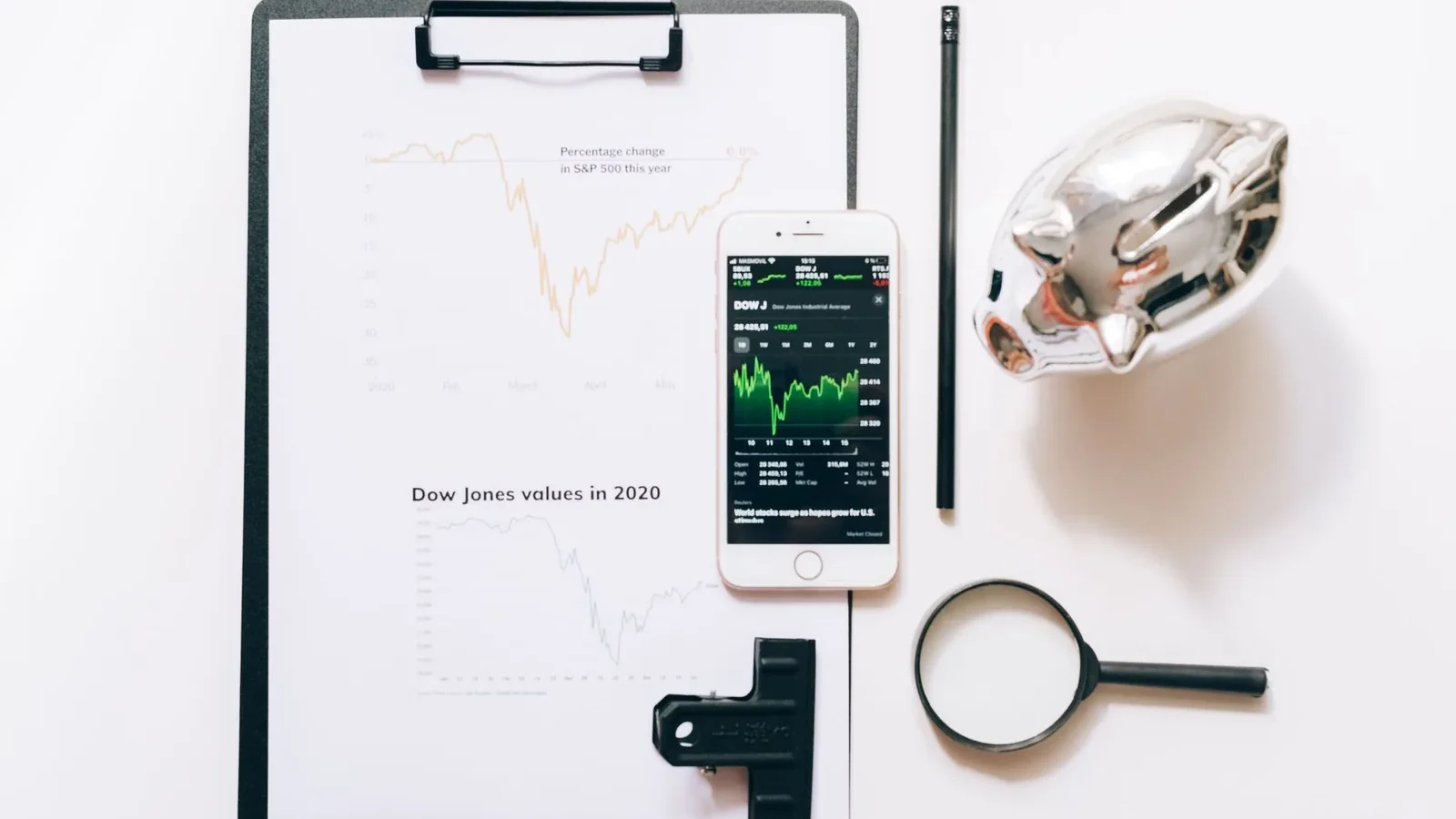 Top view of financial analysis tools including a cellphone, clipboard, magnifying glass, and piggy bank on a white desk.