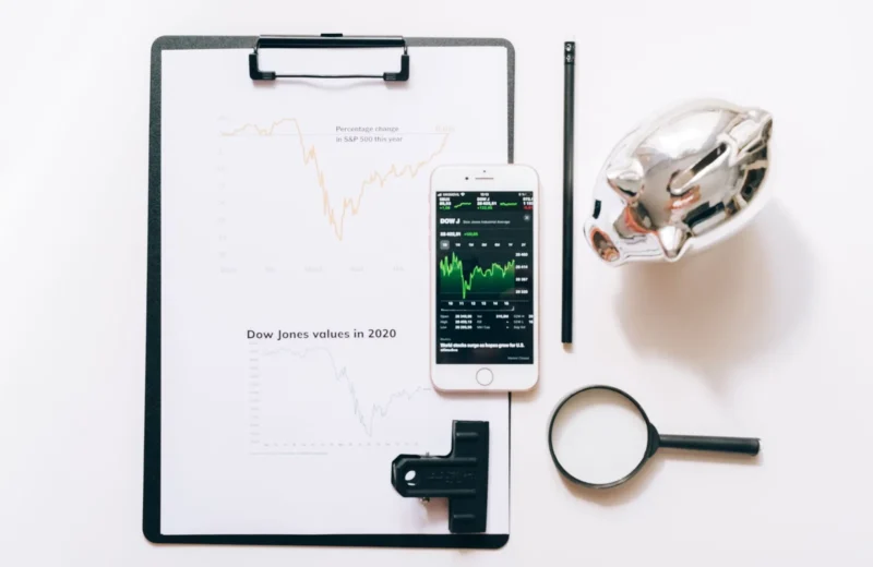 Top view of financial analysis tools including a cellphone, clipboard, magnifying glass, and piggy bank on a white desk.