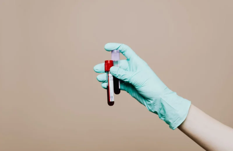 Close-up of a gloved hand holding blood test tubes against neutral background.