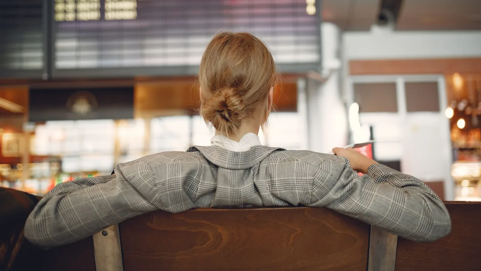 Back view of female employee in trendy jacket waiting for train departure and leaning on backs of wooden seats in train station.