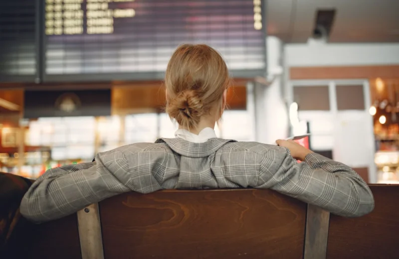 Back view of female employee in trendy jacket waiting for train departure and leaning on backs of wooden seats in train station.