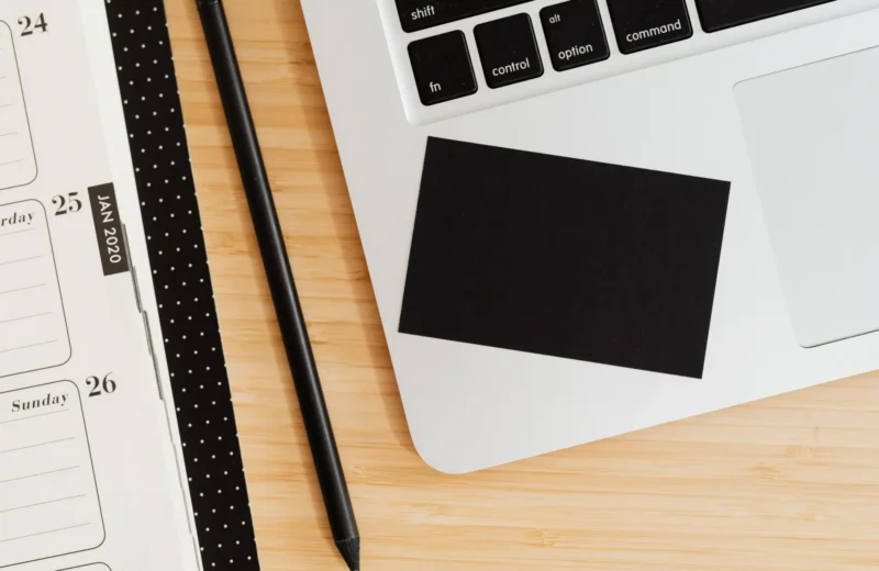 A clean office desk setup with a laptop, notebook, pen, and business card on a wooden surface.